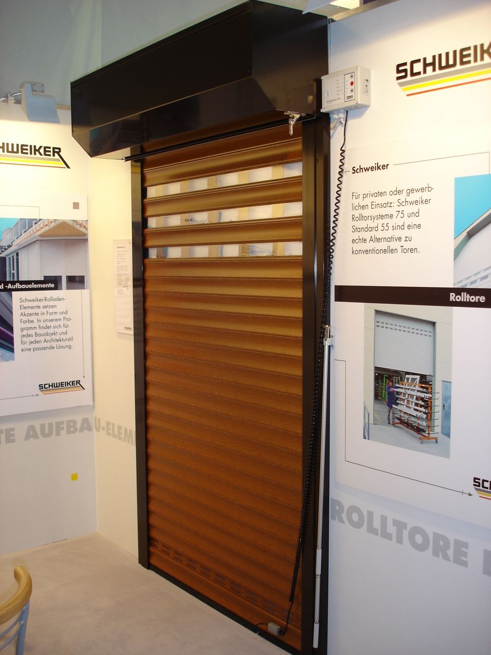 Ekspozycja bramy rolowanej w kolorze drewna z czarną obudową i sterowaniem elektrycznym, prezentowana w salonie wystawowym firmy Schweiker, z widocznymi materiałami reklamowymi na ścianie.